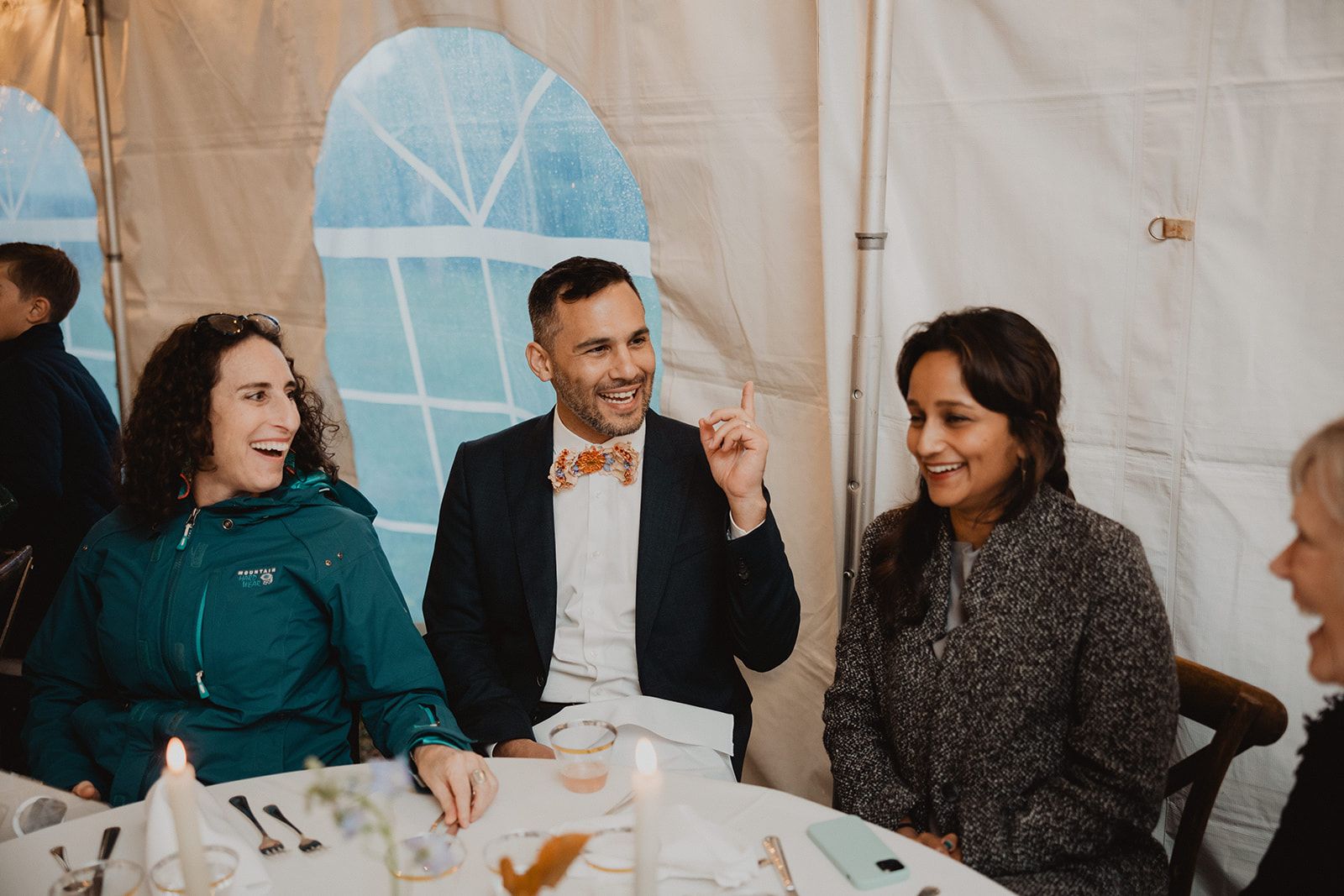 Three guests at a dinner table laugh together, the man between the two women raising a finger mid story above his patterned bow tie.
