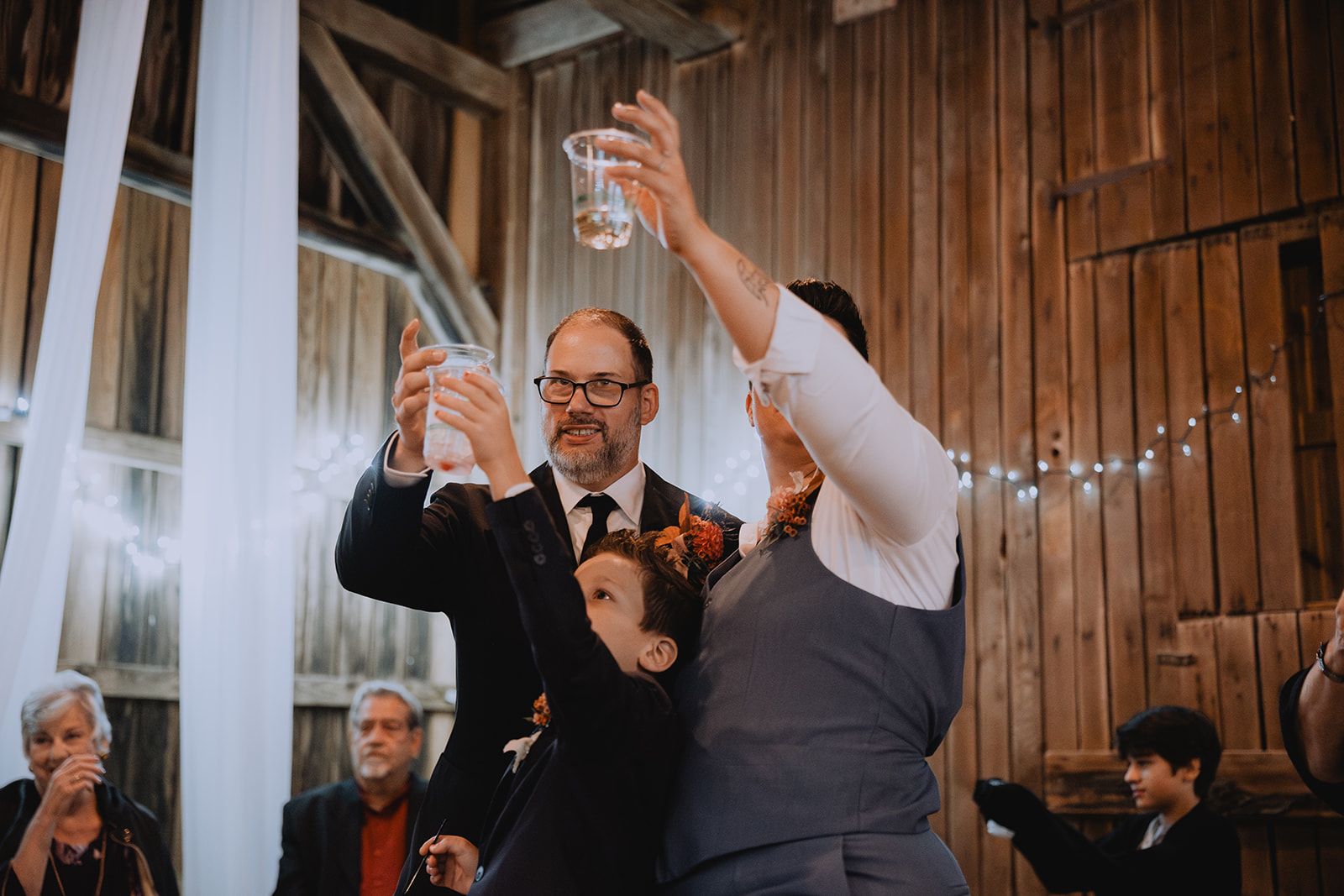 Clear plastic cups go up in a toast over a bearded man in glasses, a boy looking on from between the raised arms.