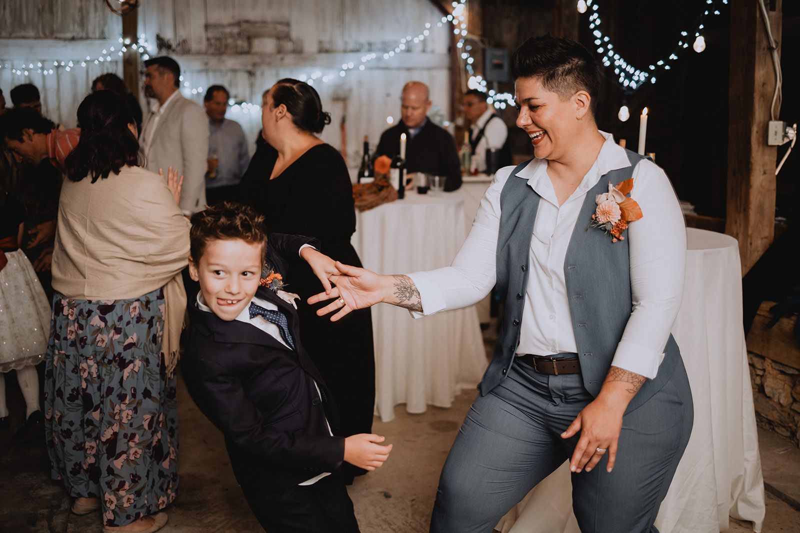 A boy in a navy suit dances with a wide grin toward the camera while a laughing adult in a gray vest reaches an arm out to him.