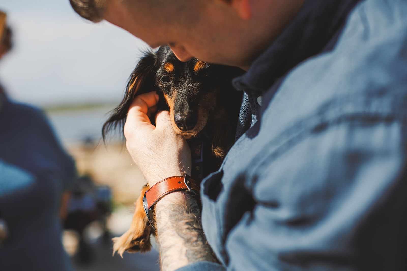 A man in a chambray shirt holds a long haired black and tan dachshund up against his chest and presses a kiss to the top of its head.
