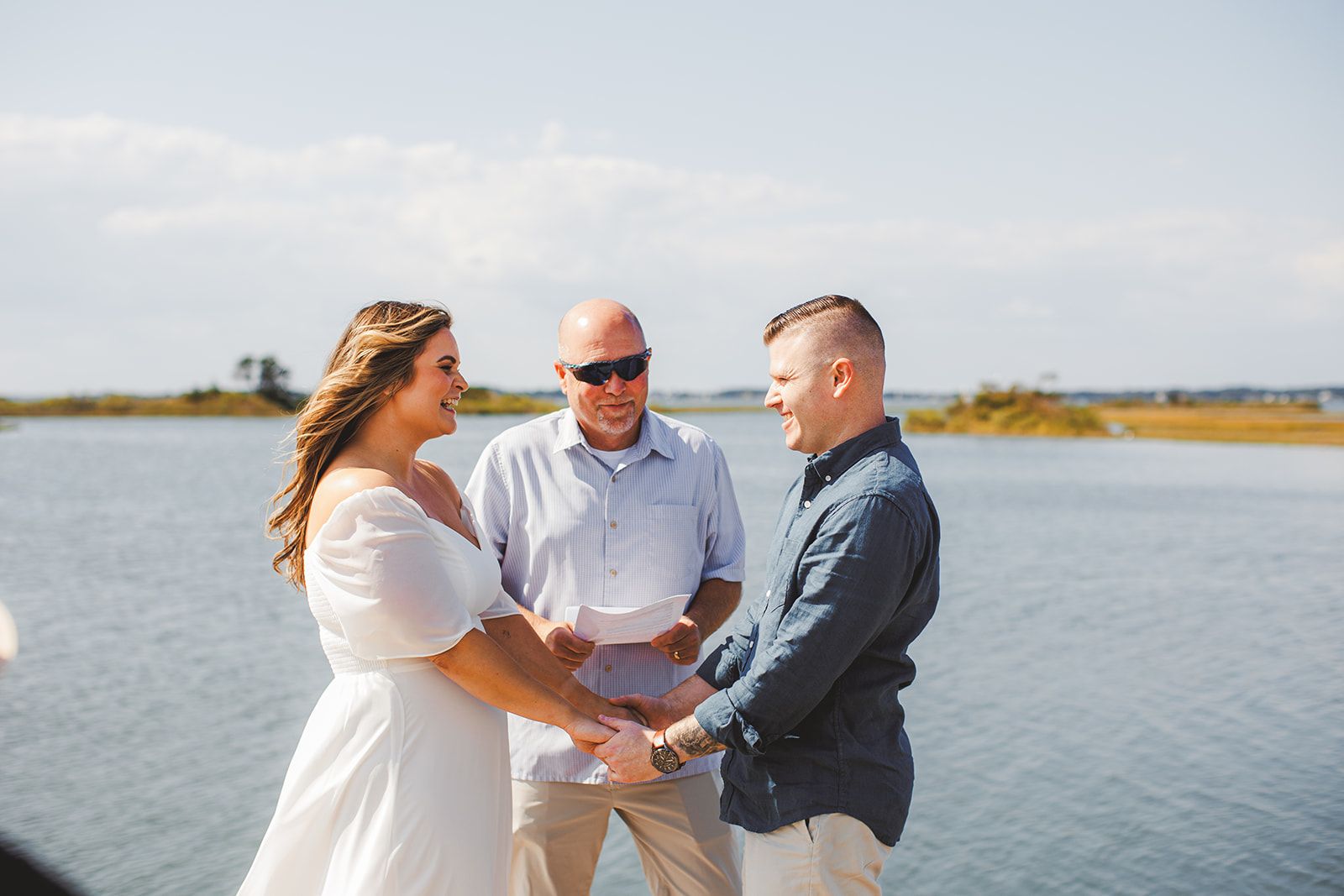 A couple hold both of each other's hands and smile at one another while a man in sunglasses stands between them holding a single page, water behind them.