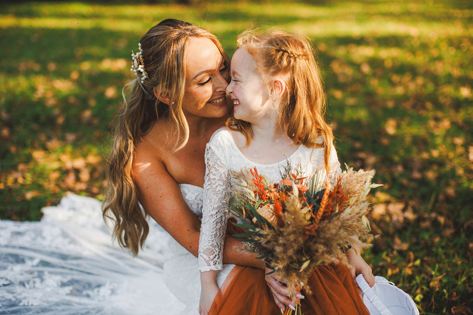 The bride crouches low in the sunlit grass with her face pressed nose to nose against a beaming young girl who holds a dried pampas bouquet.