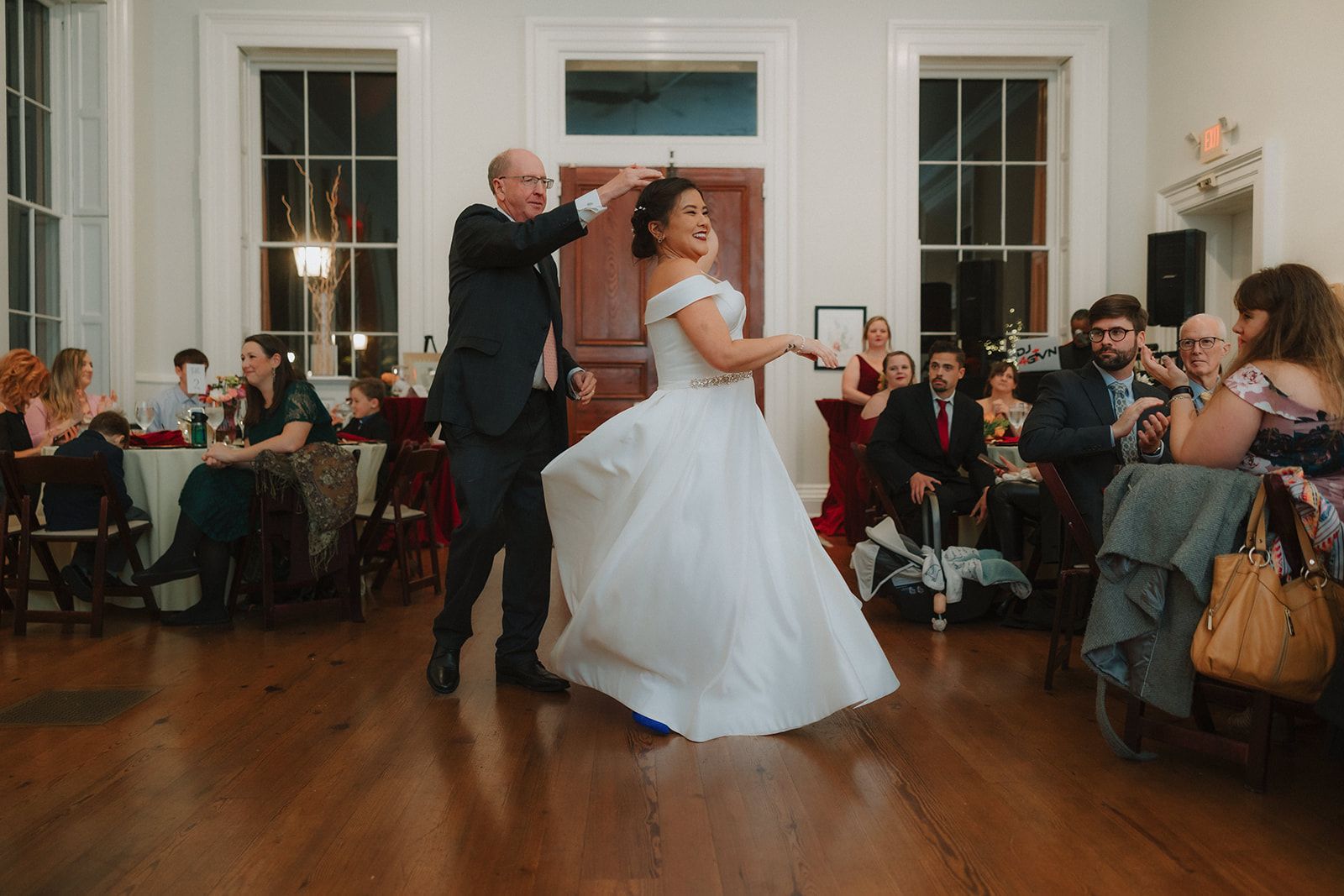 An older man in a dark suit turns the bride under his raised arm on the wood dance floor, her full skirt flaring out, as guests seated at the tables behind them look on.
