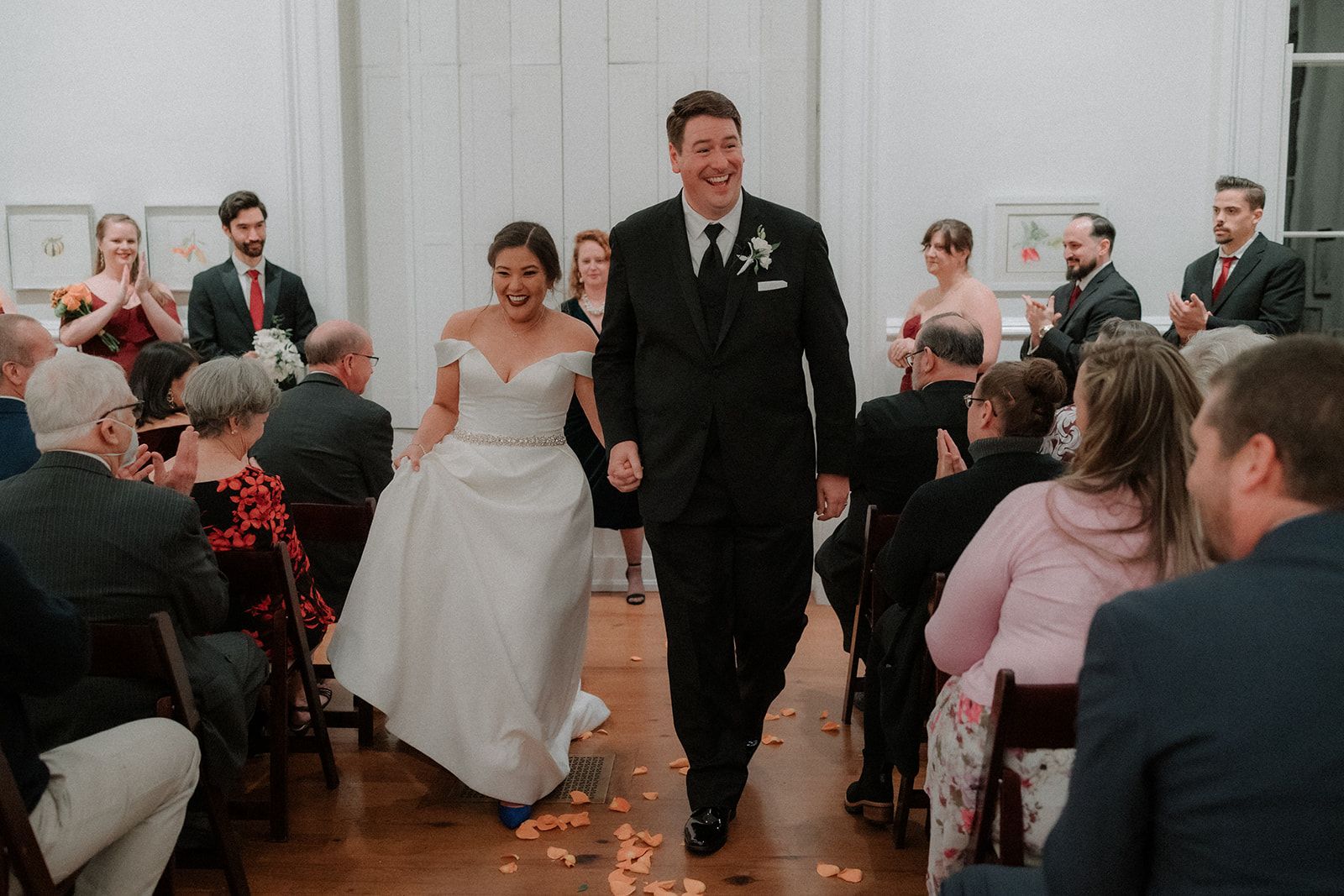 A bride and groom walk hand in hand down the petal-strewn aisle, both grinning, as seated guests turn to watch them pass.