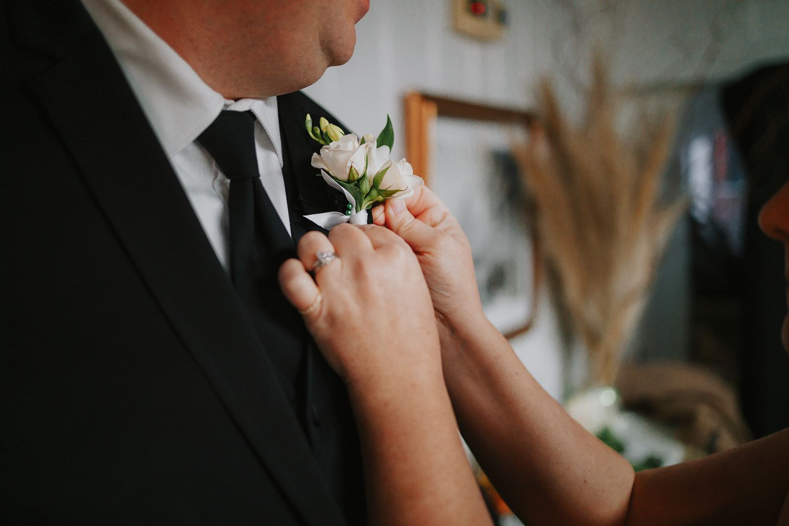 Two hands pin a white rose boutonniere to the lapel of a man's black jacket.