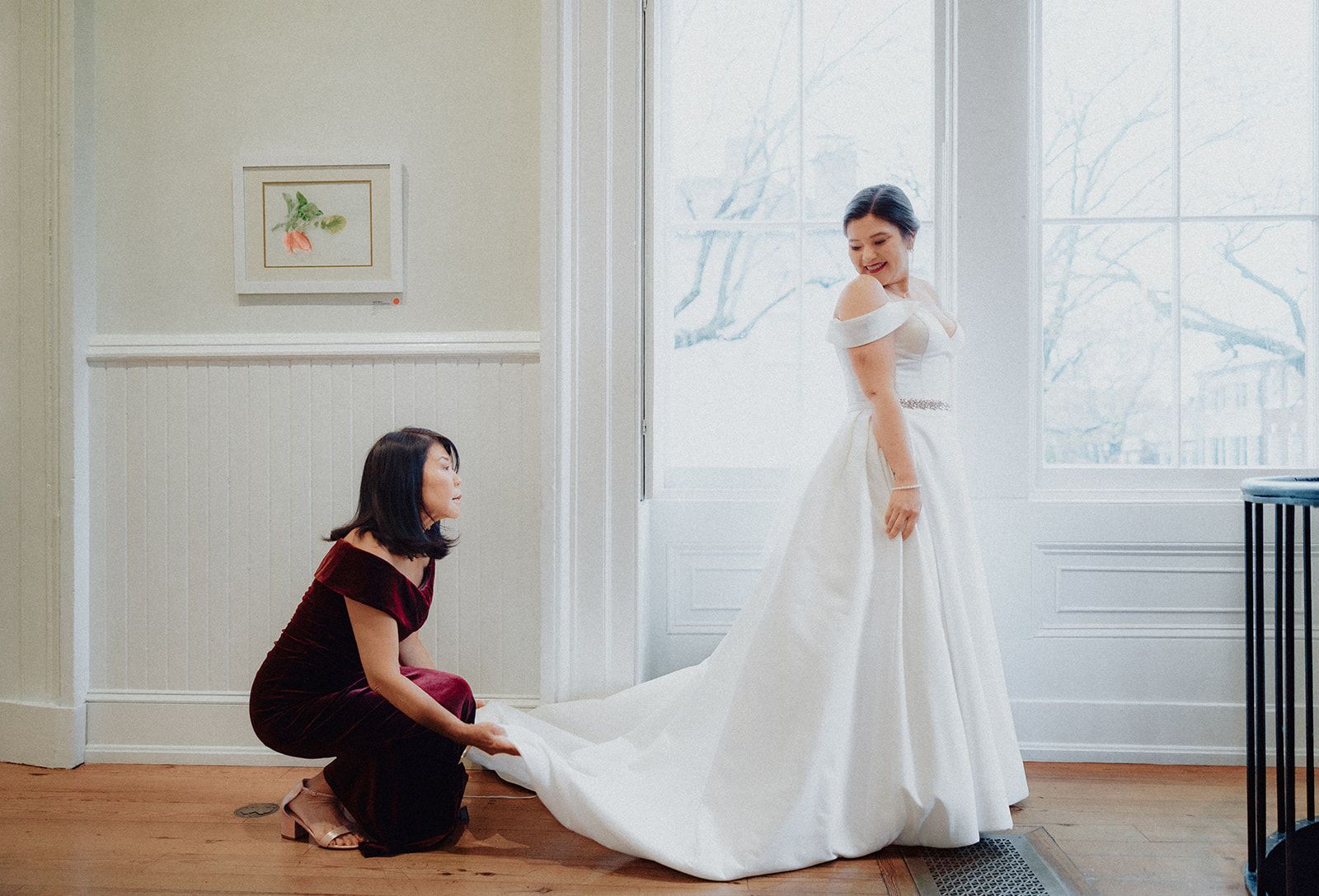A woman in a burgundy dress crouches to spread the bride's train across the floor by a bright window while the bride looks back over her shoulder.