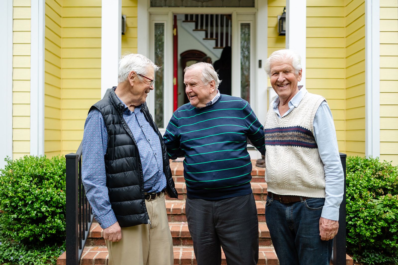 Three older men in vests and a striped sweater stand shoulder to shoulder on the brick front steps of a yellow house, laughing together.