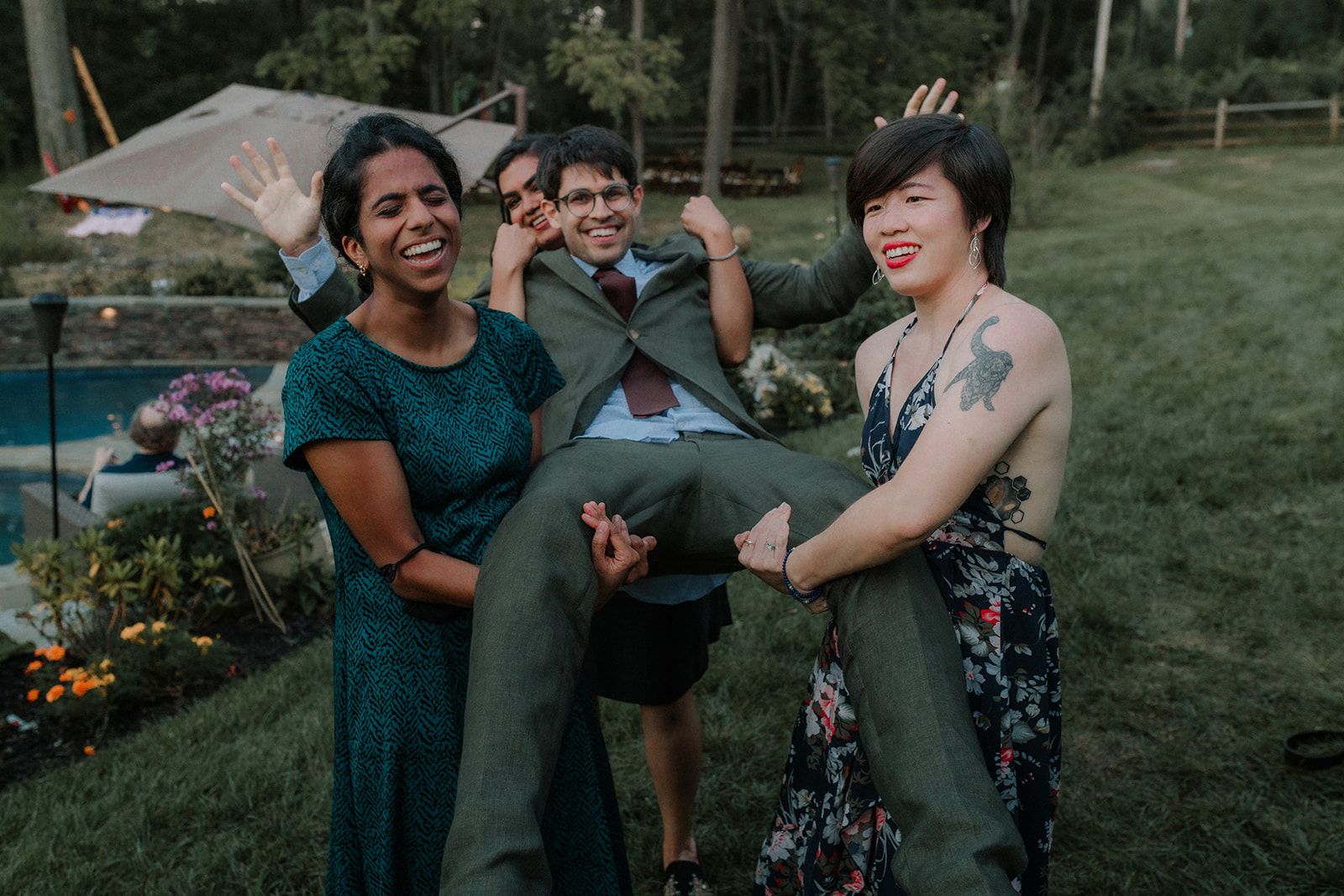 Two women hoist a grinning man in a green suit off the ground while he flexes an arm and another guest laughs over his shoulder behind them.