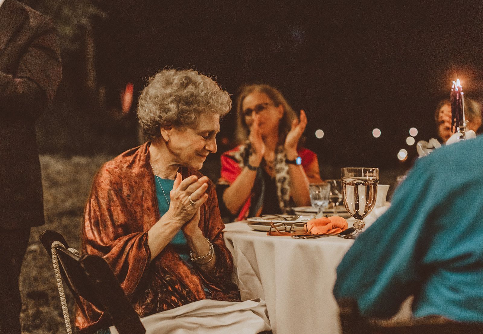 An older woman in an orange wrap claps and smiles at a candlelit table while a woman in glasses applauds just behind her.