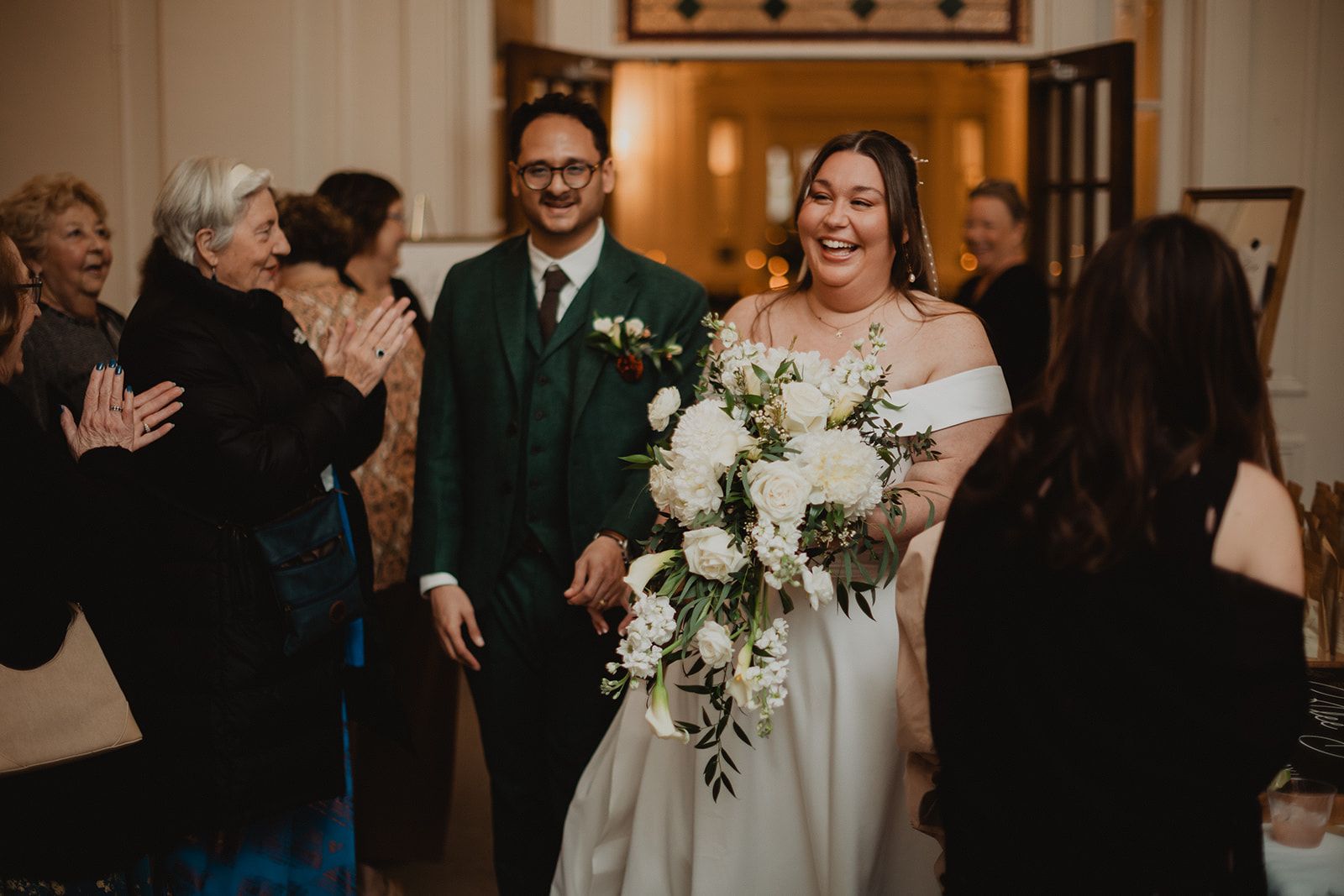A row of guests clap and smile as the bride walks in with a large white bouquet beside a man in a green suit.