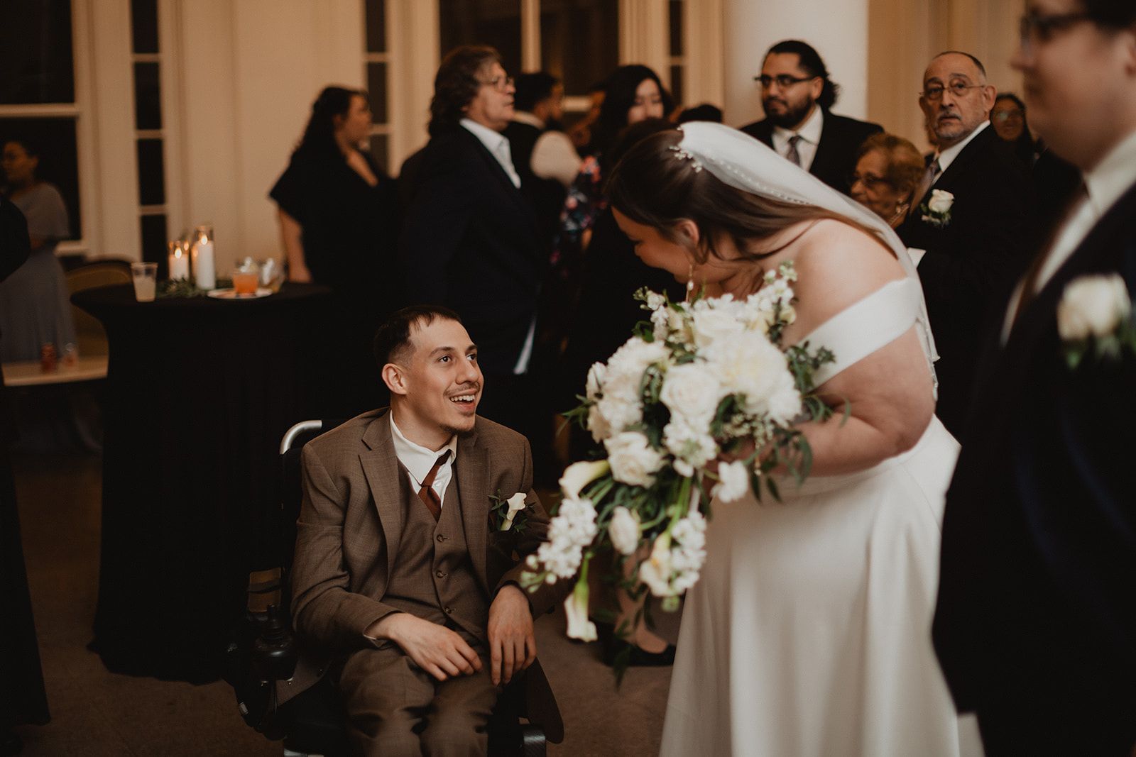 A young man in a brown three piece suit seated in a wheelchair grins up at the bride as she leans down toward him with her bouquet.