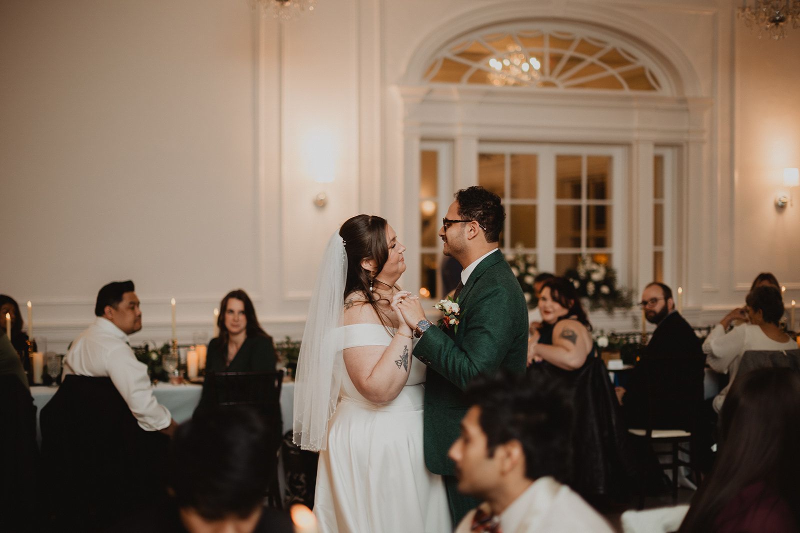 The groom cups the bride's cheek as they dance close together, their other hands clasped between them, seated guests all around.