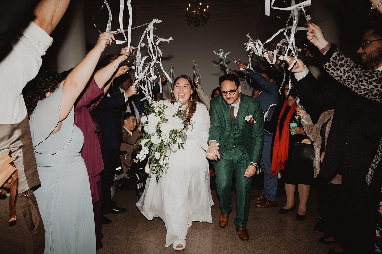 Guests raise white ribbon wands overhead on both sides as the couple walks through hand in hand, the bride laughing.