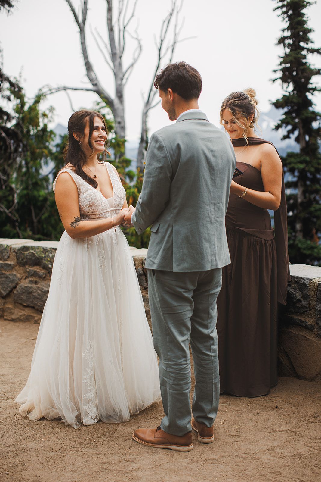 The bride smiles at the groom as they hold hands during the ceremony while a woman in a brown dress stands beside them with her eyes lowered.
