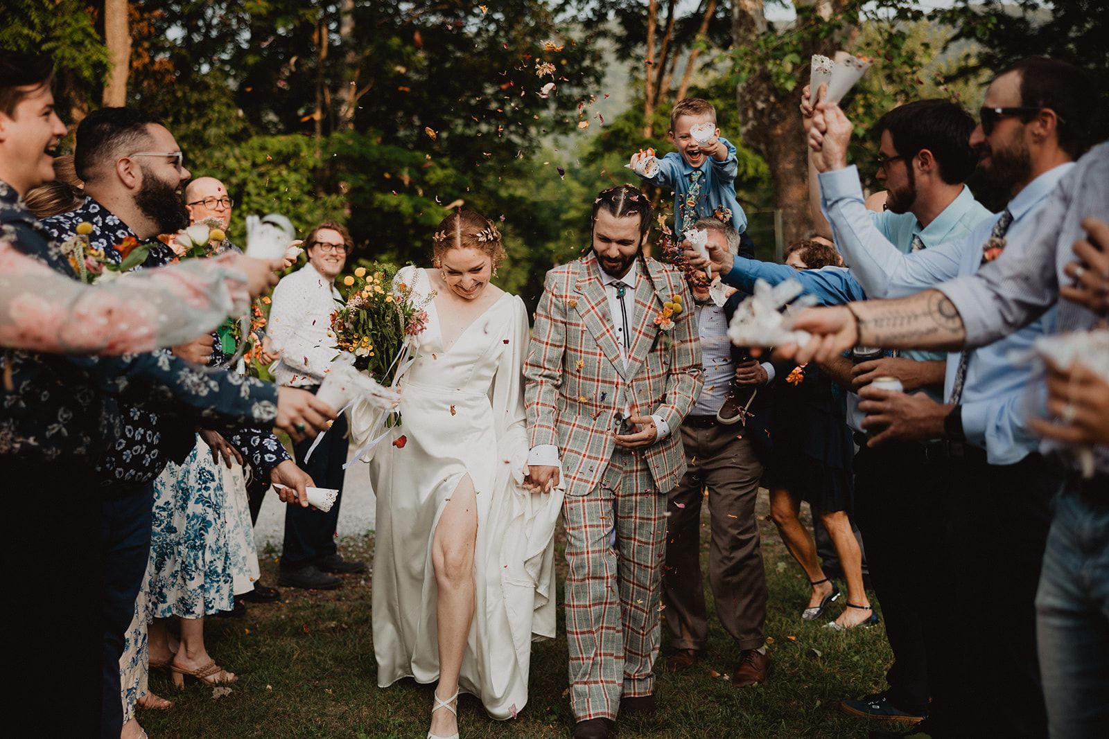 Guests line both sides of a grass path tossing petals as the couple walks through hand in hand, with a small child riding on someone's shoulders above the crowd.