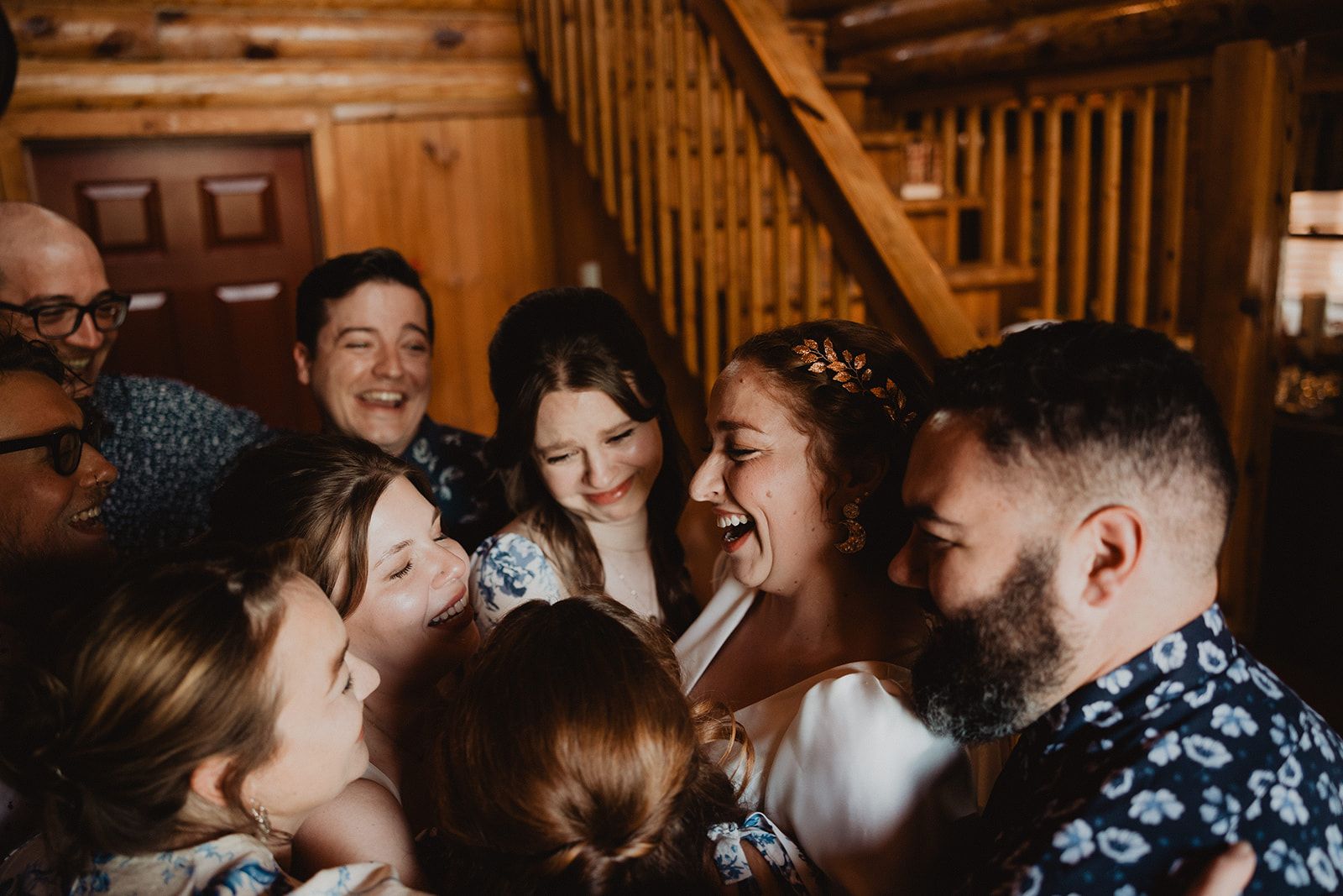 A tight huddle of people presses in around the laughing bride inside a log cabin room.