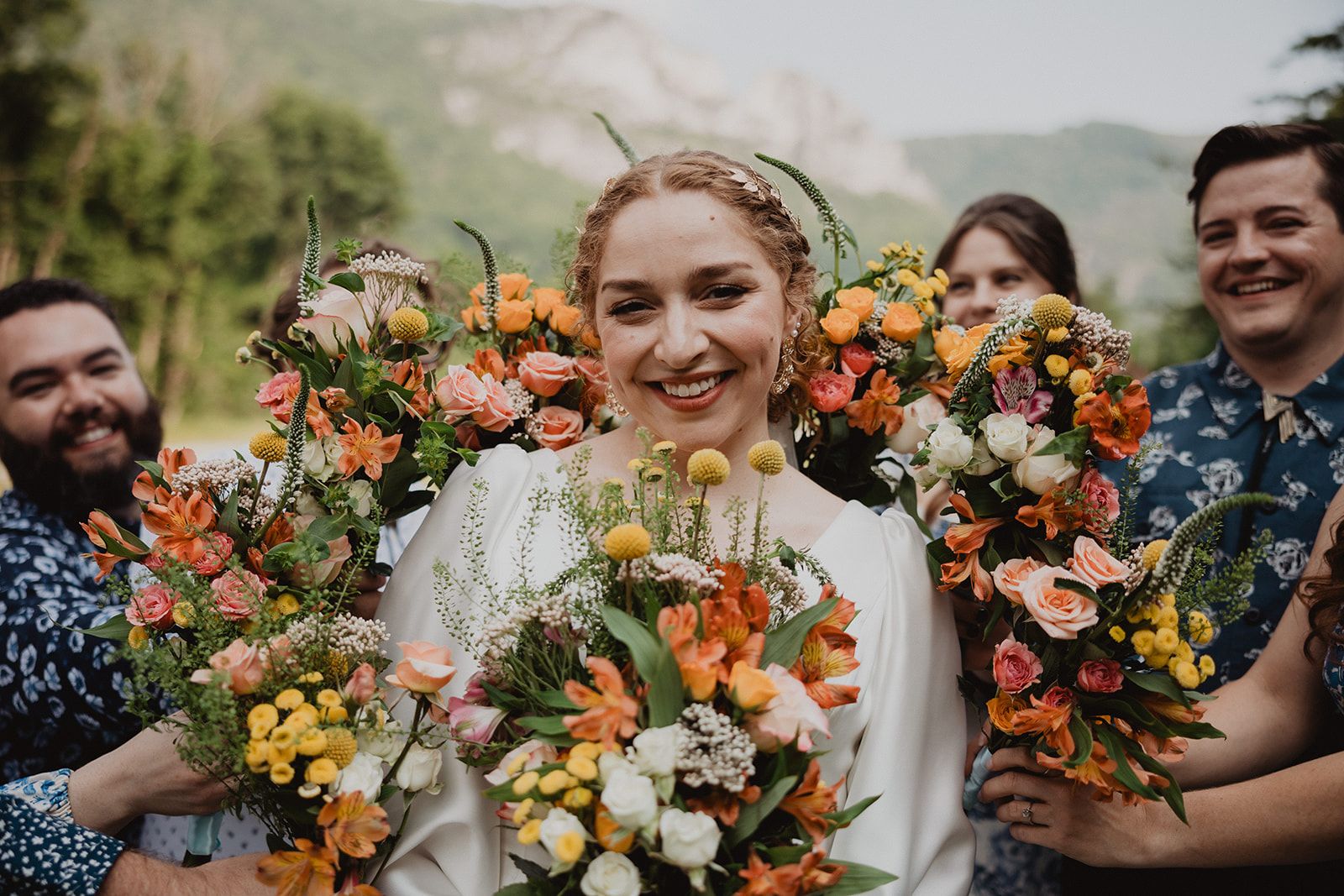 The bride grins at the camera while the smiling people crowded around her hold bright orange and yellow bouquets up on either side of her.