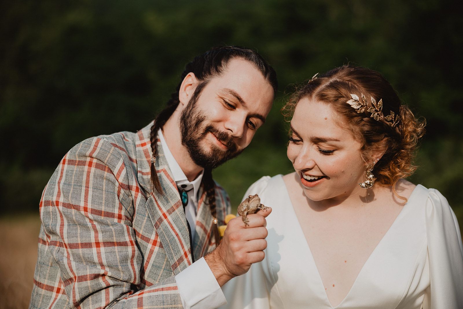 The groom holds a small frog on his fingers while the bride laughs beside him, both looking down at it.