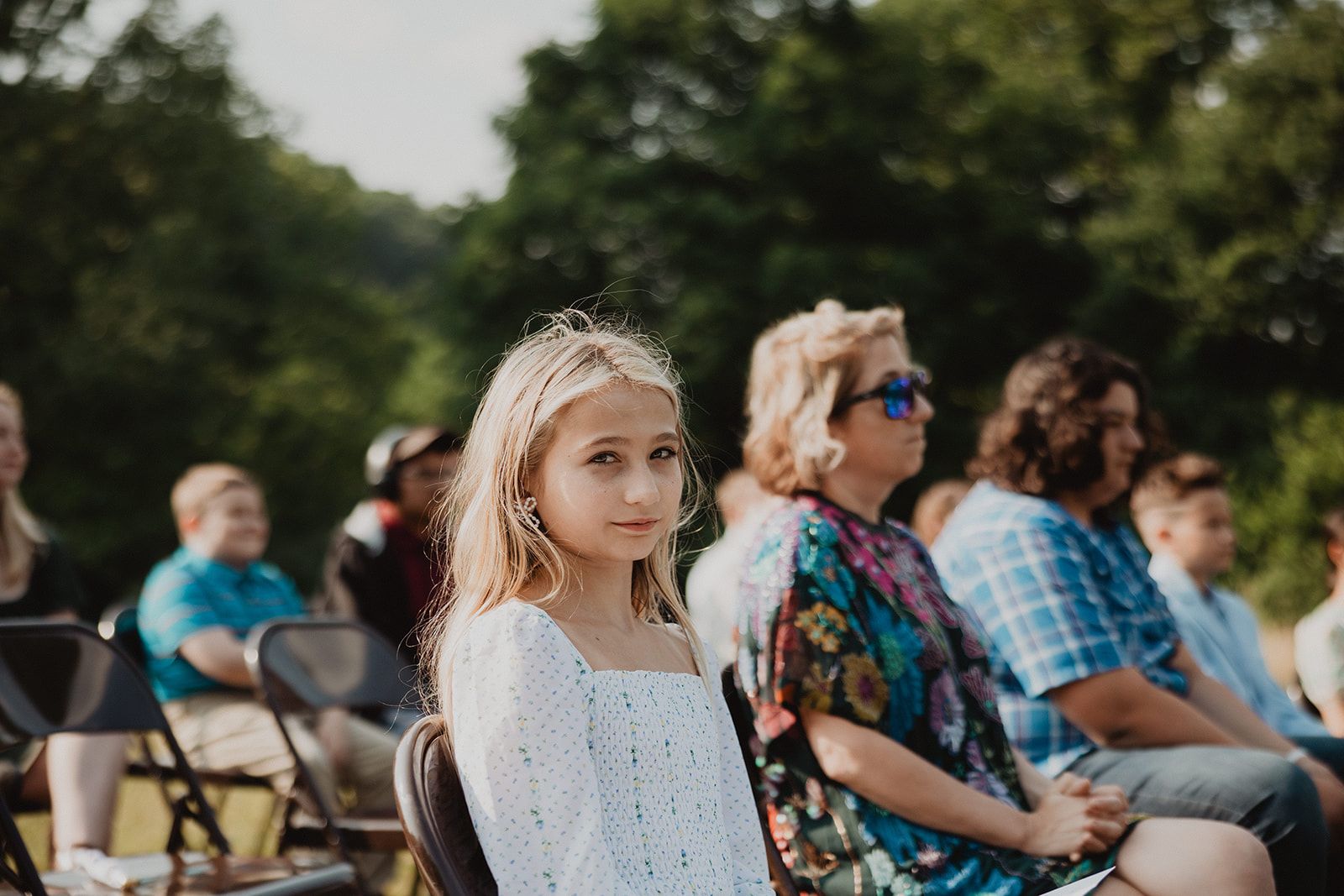 A young girl in a white dress sits among rows of seated guests outdoors and turns to look back toward the camera.