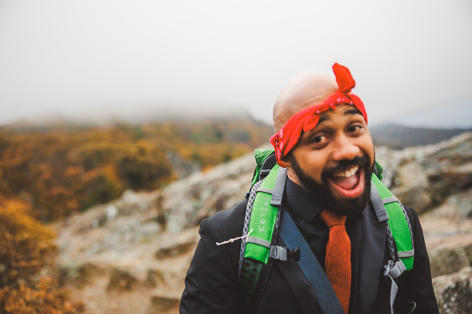 A man in a suit and red bandana with a green pack on his back laughs open mouthed on a fog covered hillside of autumn brush.