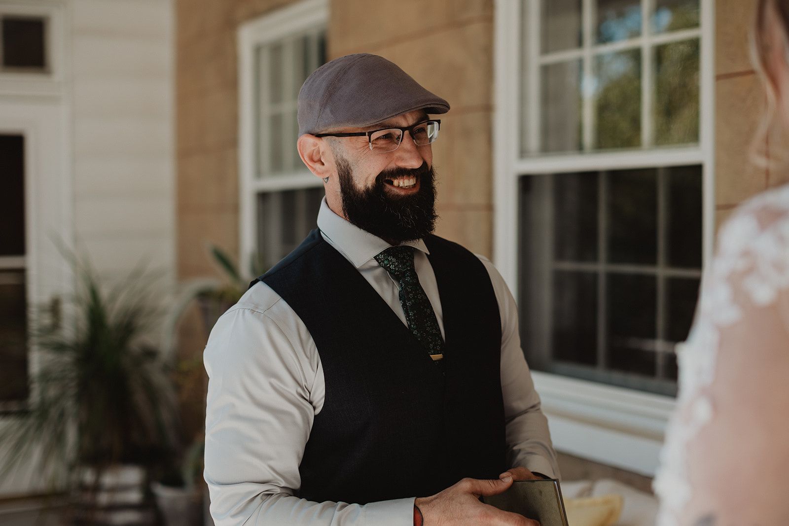 A bearded man in a flat cap, glasses and a black vest grins toward a blurred figure in a lace gown at the right edge of the frame.