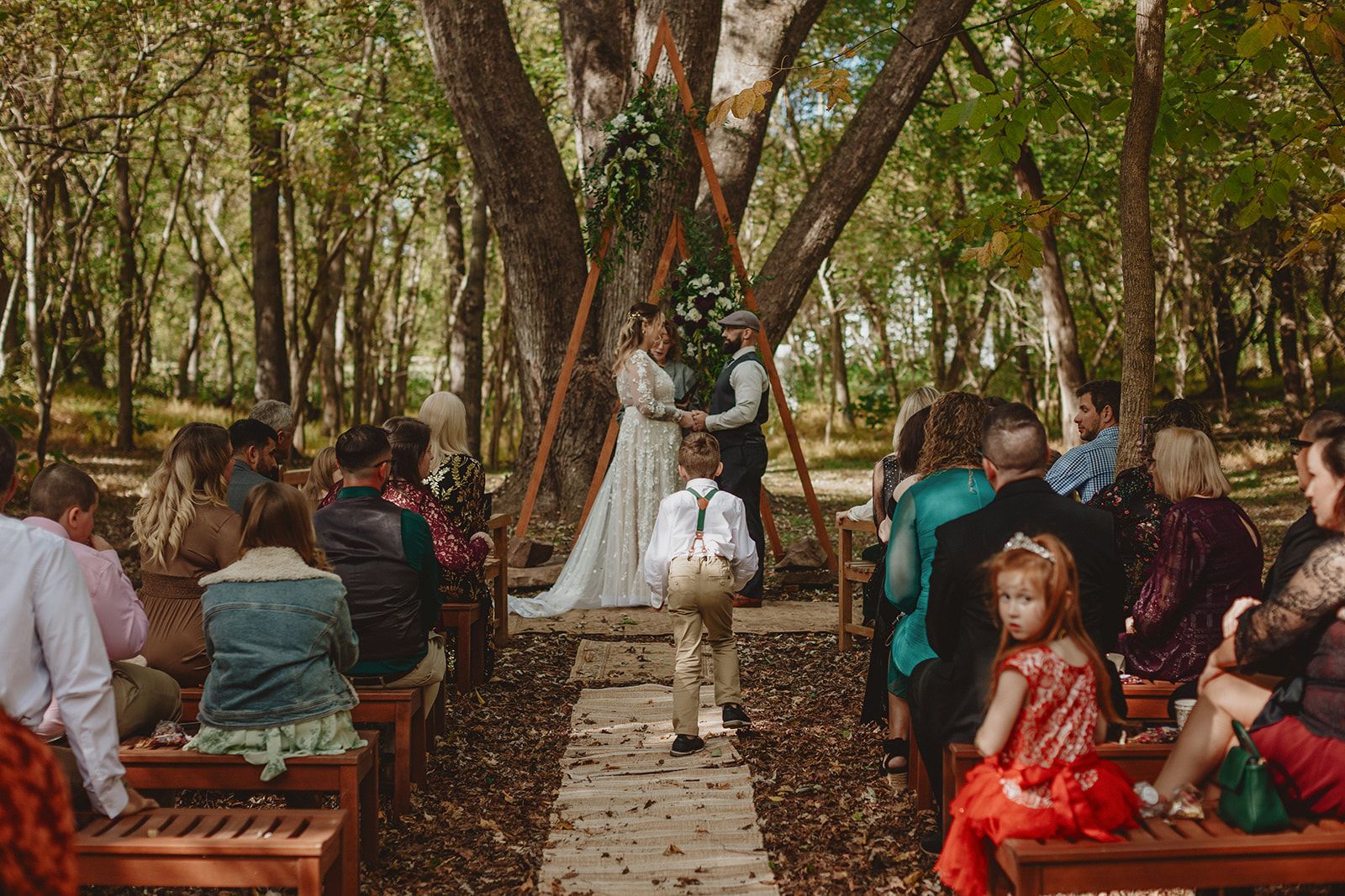 A boy in green suspenders walks alone up a wooden plank aisle between benches of seated guests toward the couple standing hand in hand under a flowered wooden arch.