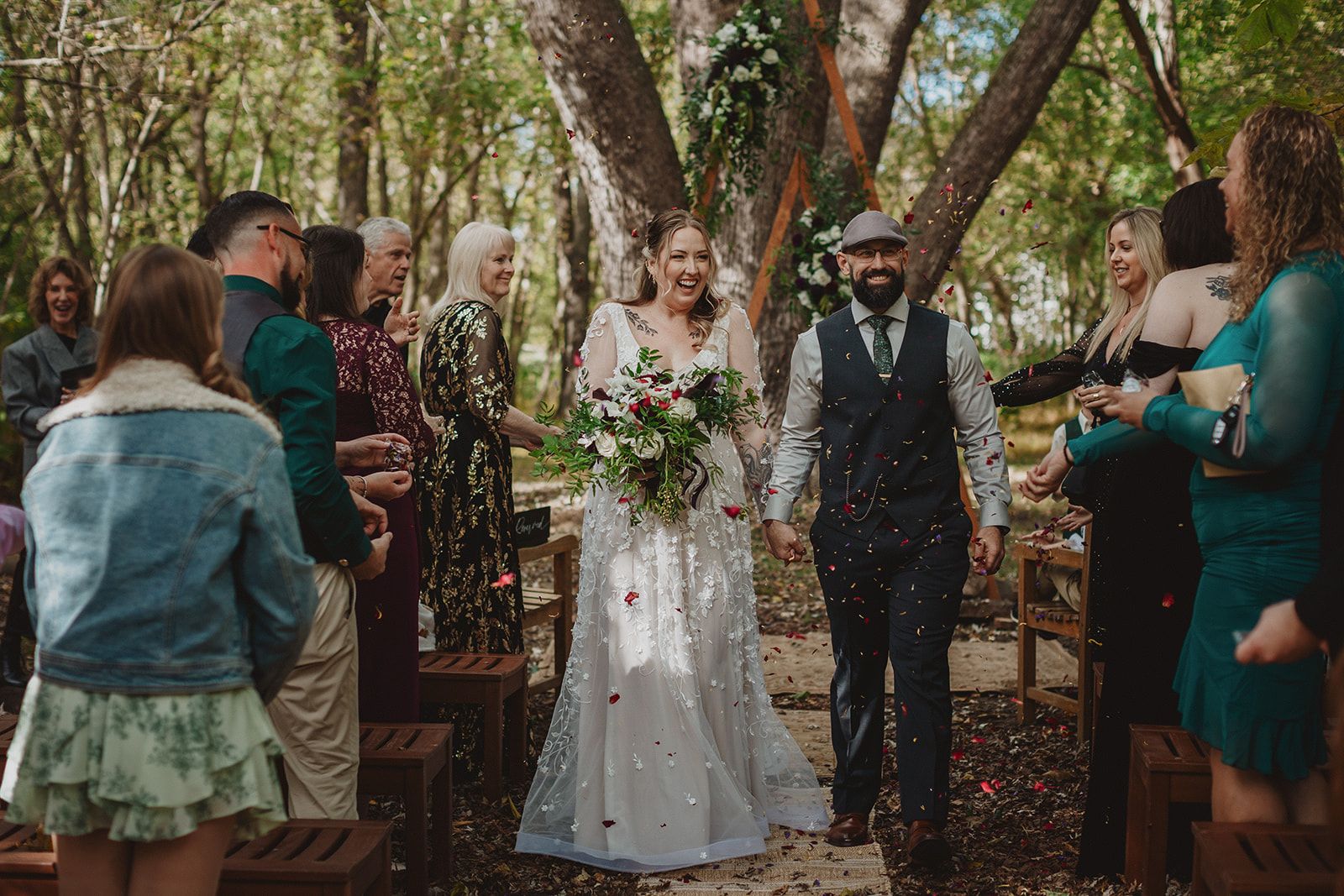 The bride beams with her bouquet in one hand and the groom's hand in the other as they walk the aisle between standing guests through falling confetti.