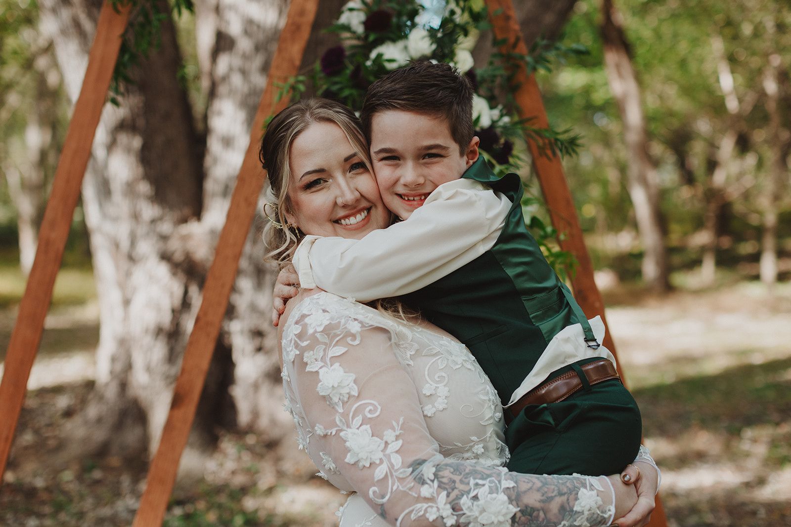 A woman in a white lace gown grins while a boy in a dark green suit hugs her tightly around the neck under a flower covered wooden arch.
