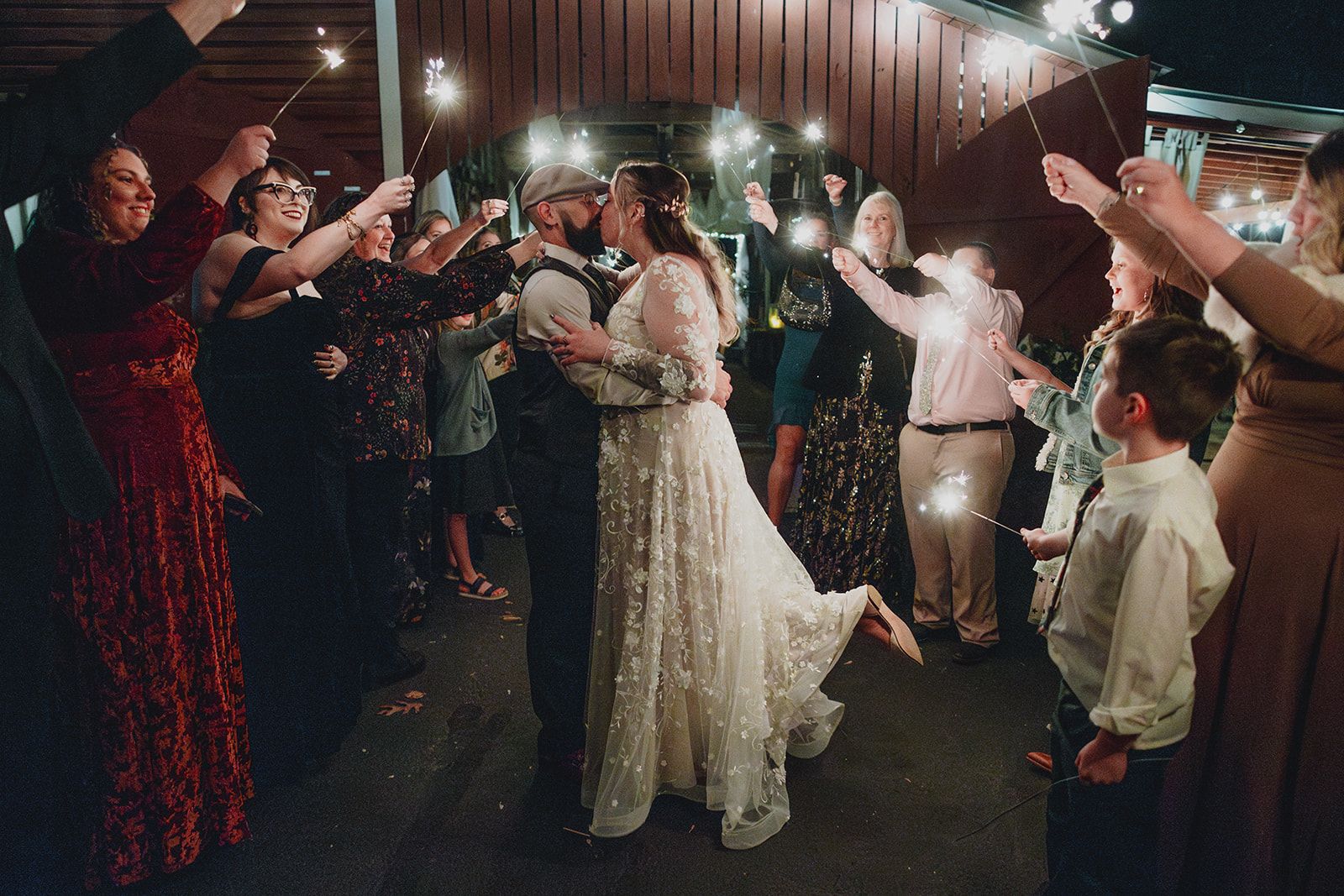 Guests raise lit sparklers in two lines at night as the couple kisses between them and a boy in the foreground watches with a sparkler in his hand.