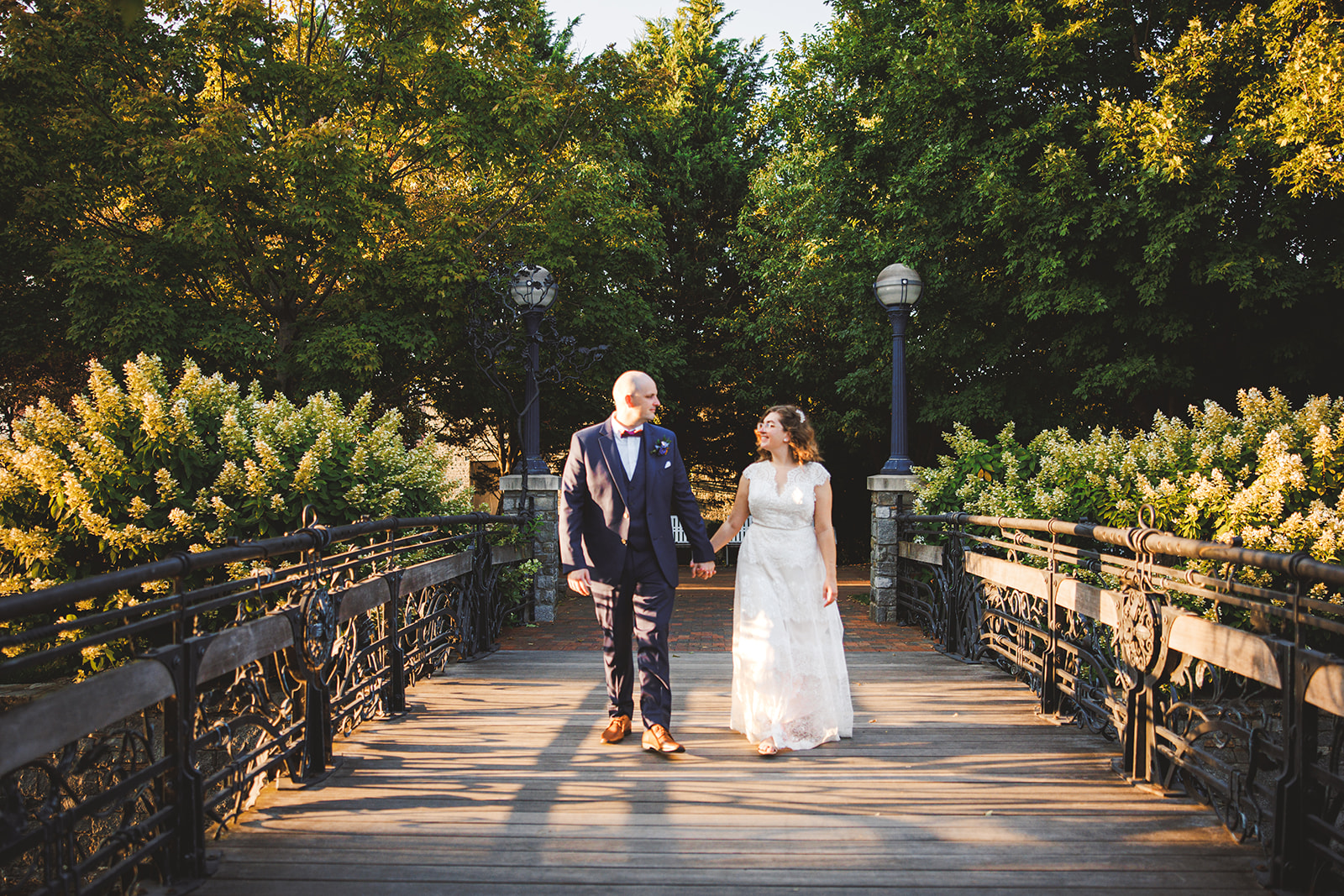 A couple walking hand in hand across an arched bridge over Carroll Creek in downtown Frederick, iron railings either side and trees closing overhead in evening light