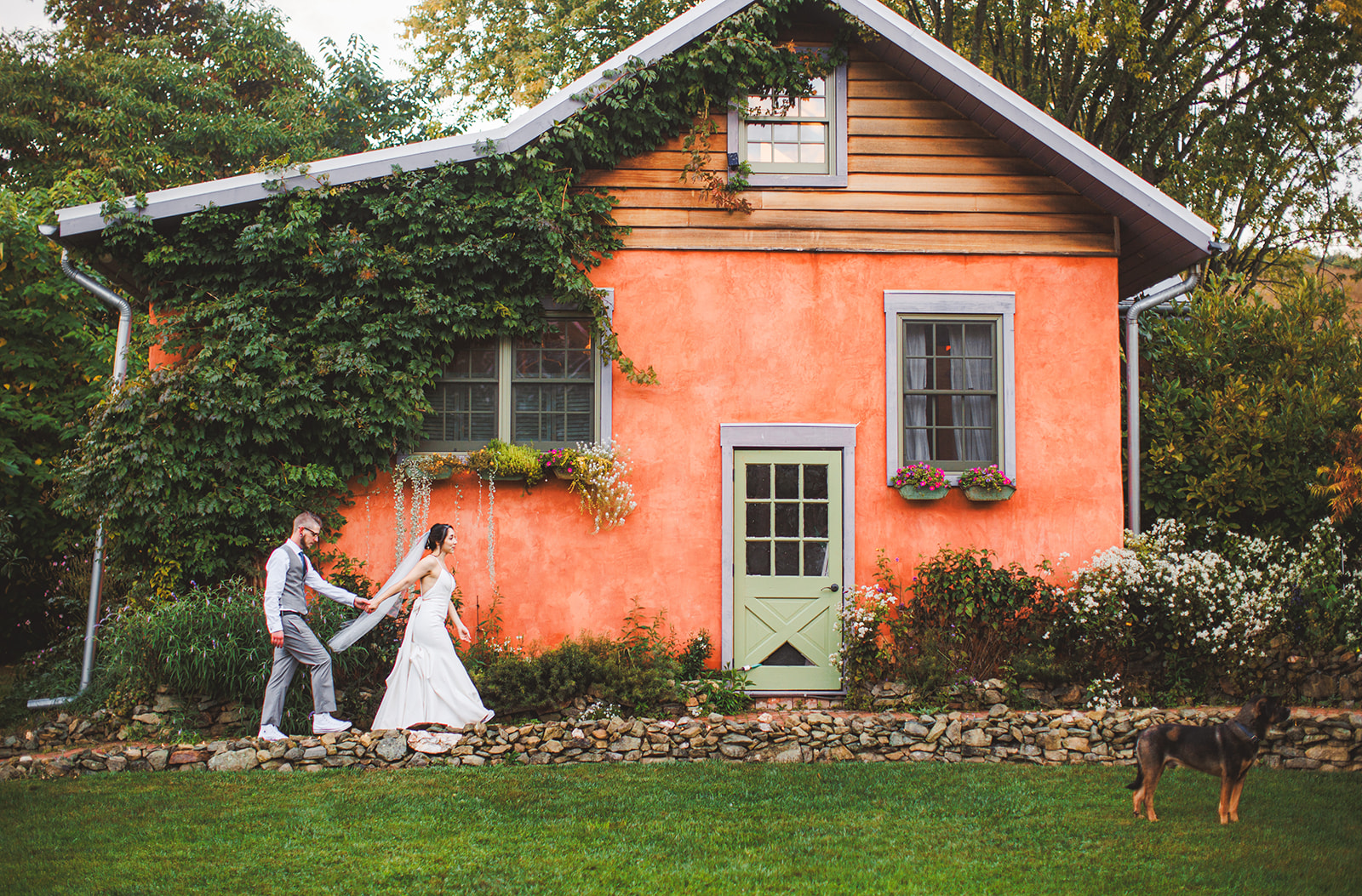 A couple walking hand in hand across the lawn in front of a coral-painted farmhouse draped in ivy, green shutters and a small porch behind them
