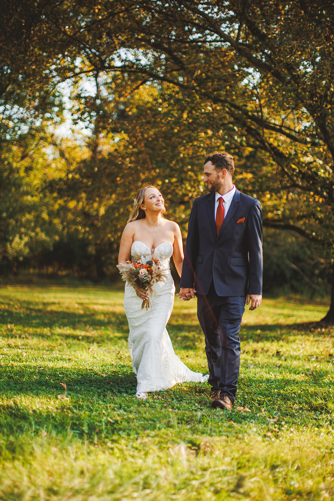 A couple walking together through a golden evening field at Fox Haven Farm, her bouquet down at her side and low sun catching the grass around them