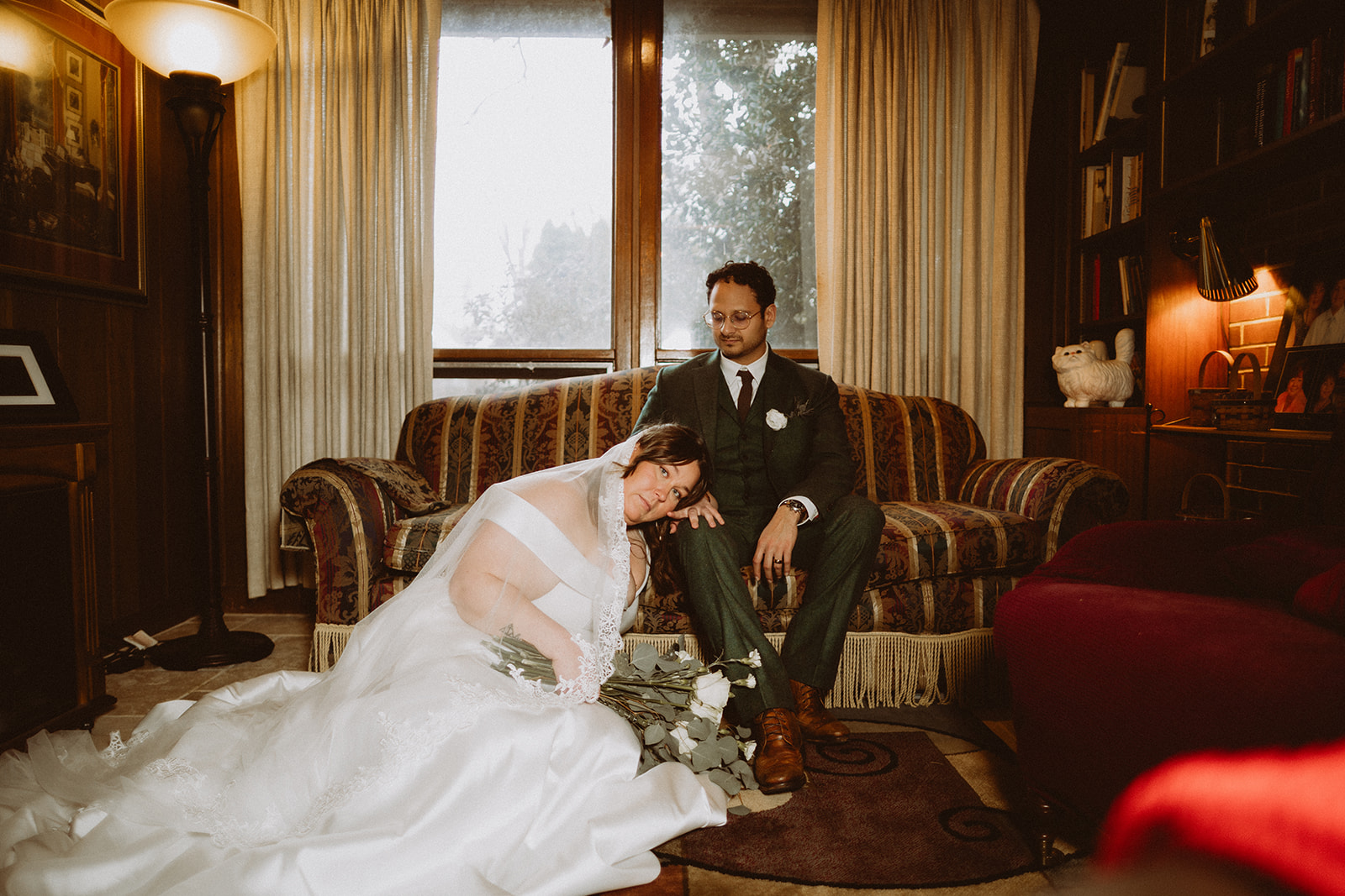 Mersades resting her head on Rogelio's shoulder on a striped sofa in a wood-panelled room, her dress spread across the floor and winter light coming through the window behind them