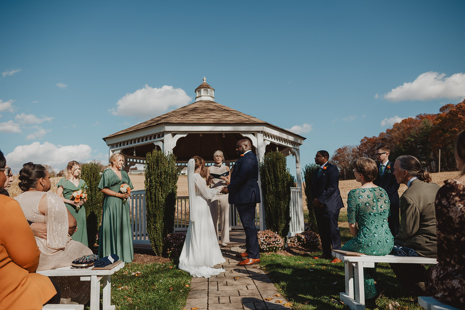 A gazebo ceremony on a bright autumn day, the couple facing each other under the open timber roof with guests seated on white benches either side