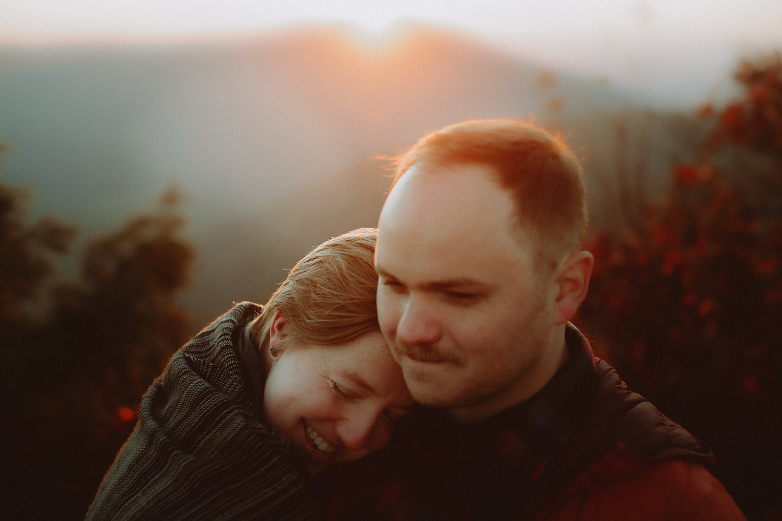 A couple wrapped together in a blanket at sunset, foreheads touching, the sun dropping behind hazy blue ridgelines and lighting the edges of their hair