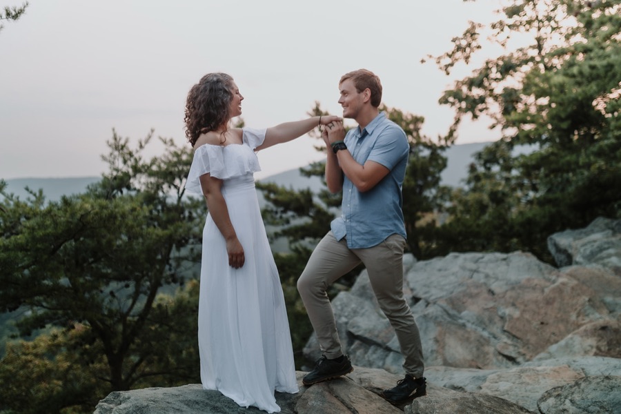 A man on the rocks lifting a woman's hand to kiss it, her white dress catching the last light, pines and a hazy river valley falling away behind them