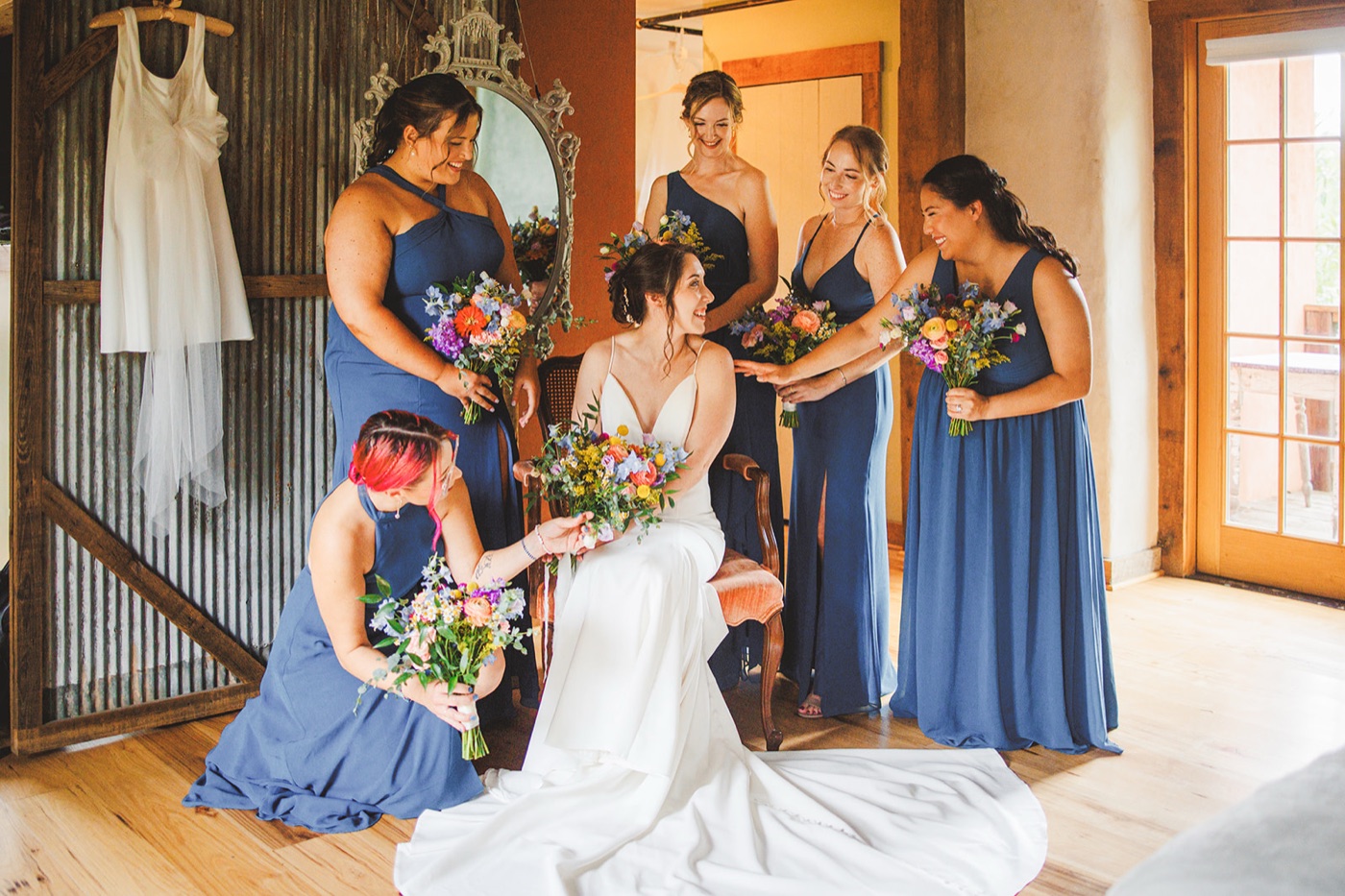 A seated bride holding a wildflower bouquet as bridesmaids gather around her, one kneeling to arrange her gown's train