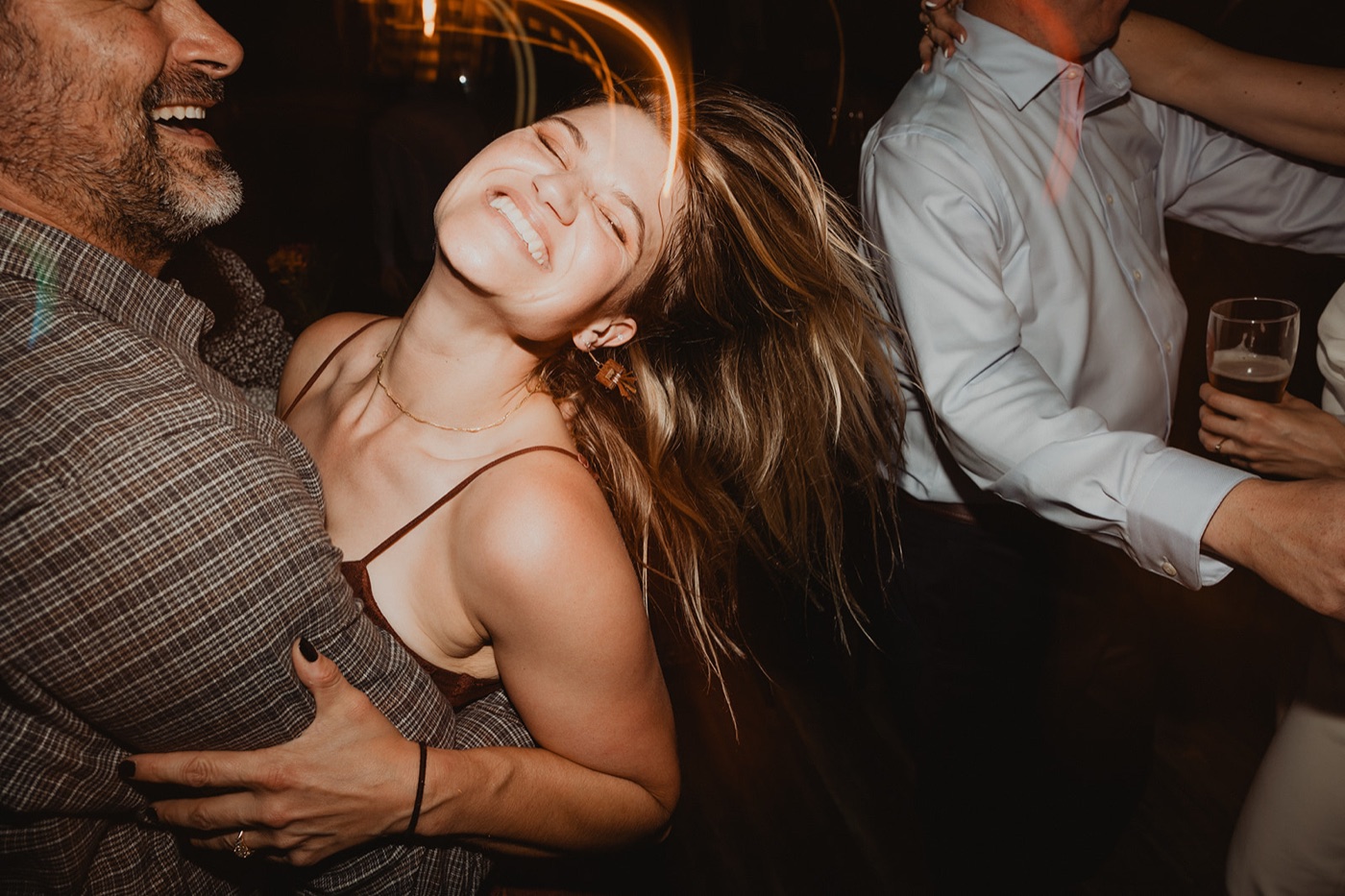 A woman throwing her head back laughing while dancing between two men at a dimly lit reception