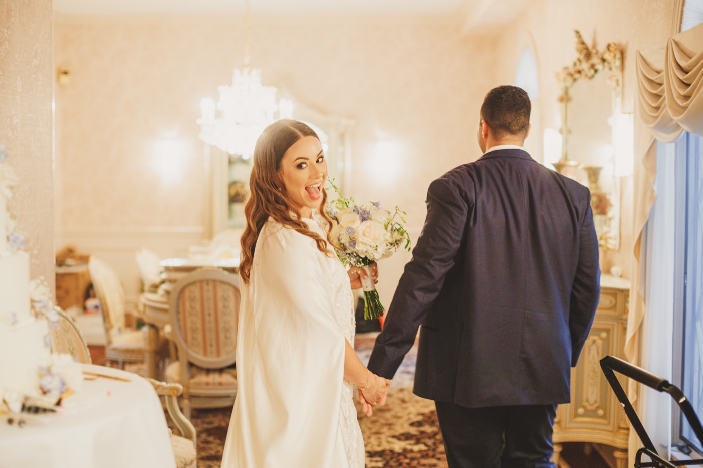 A bride glancing back with a playful open-mouthed smile while holding the groom's hand in an ornate mirrored ballroom