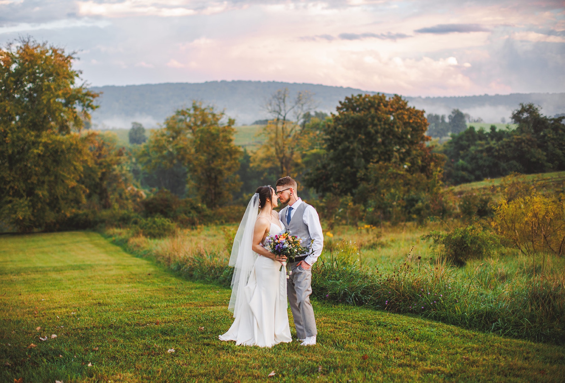 A couple forehead to forehead in a golden meadow with misty layered mountains and a pastel sky behind them